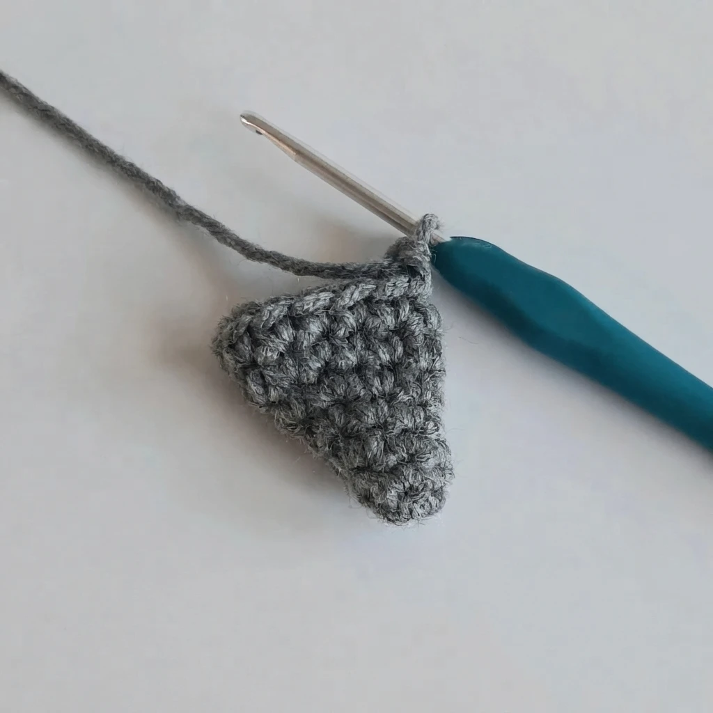 Small gray crocheted fin in progress on a plain white surface, still held on a teal-handled crochet hook, with the working yarn trailing up to the left.
