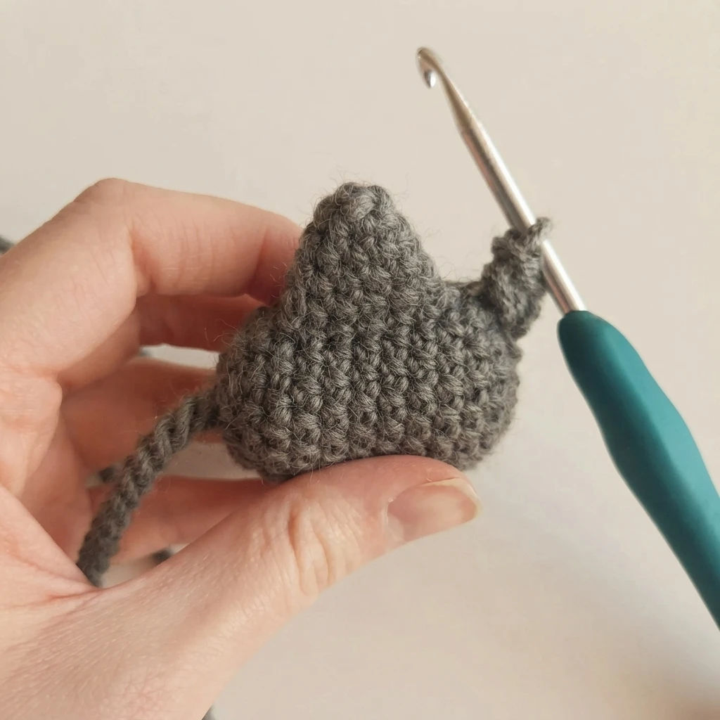 A hand holds a small gray crocheted fin in progress, worked with a teal-handled crochet hook, with a short yarn tail hanging below, against a plain light background.