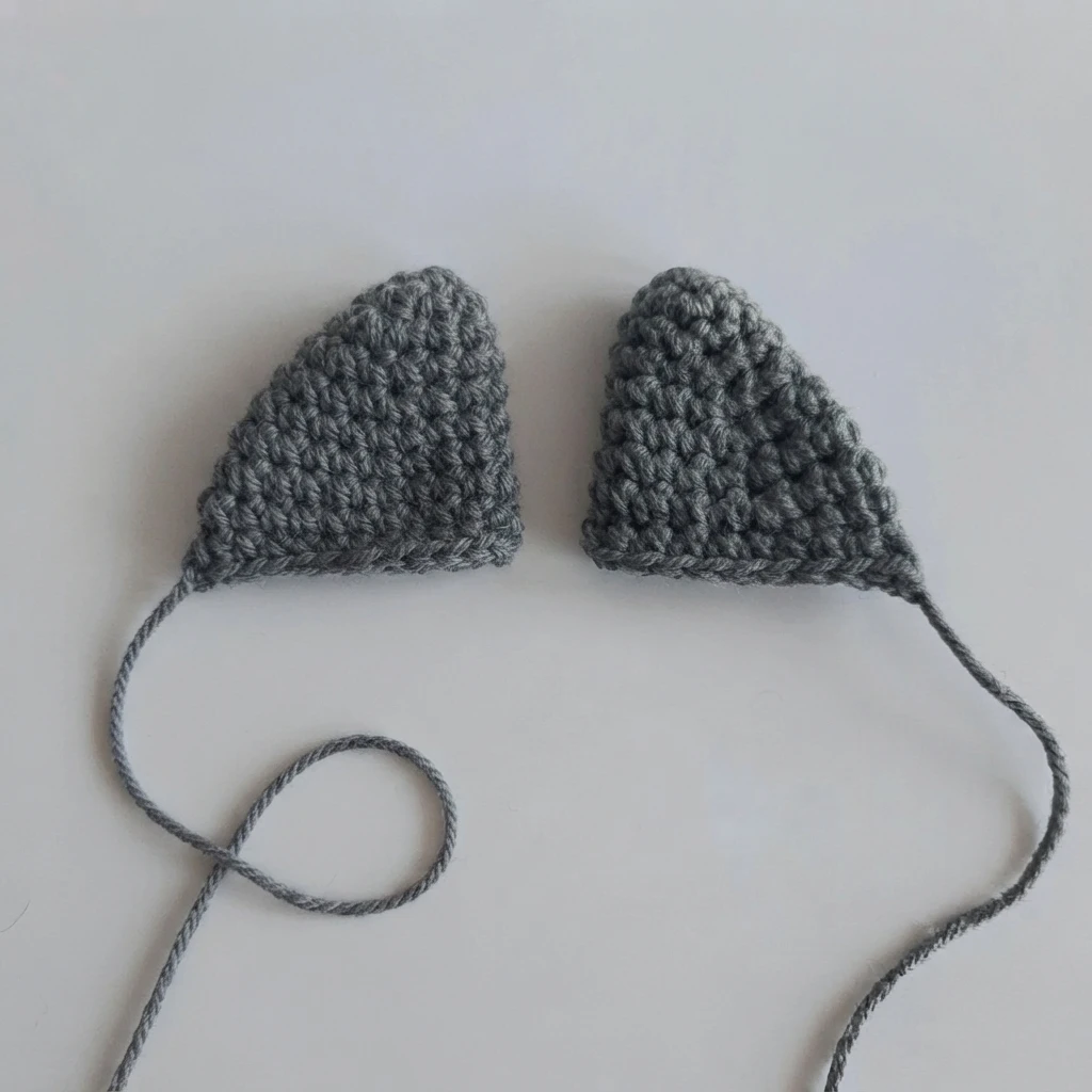 Two gray crocheted triangular fins with long yarn tails resting on a plain white surface, the left tail yarn curled into a loop.