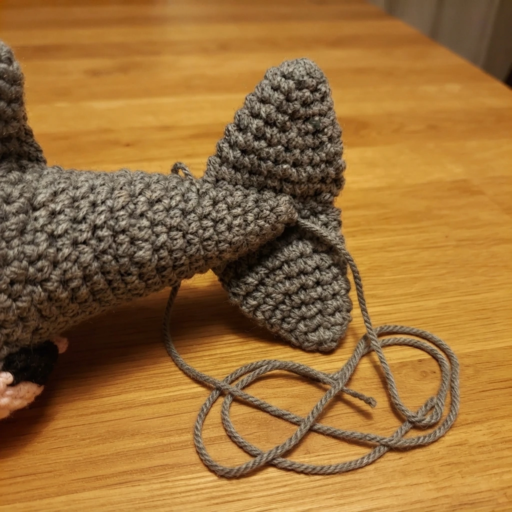 Close-up of a gray crocheted shark tail with two triangular fins sewn together and attached to the tail, loose gray yarn tails resting on a light wooden table.