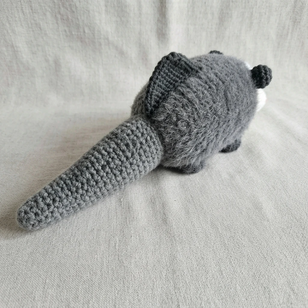 Gray crocheted possum plush seen from behind on gray fabric, with a long tapered gray tail sewn to the body, a black fin on the back and small black ears.