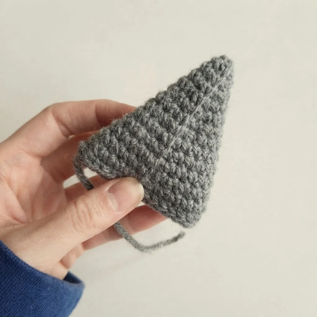 A hand holding a small gray crocheted triangular fin with a short yarn tail hanging below, photographed against a plain pale background.