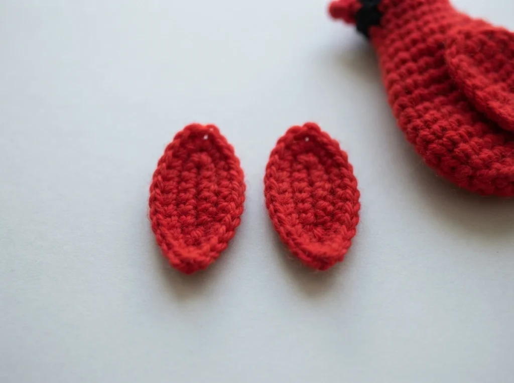 Two flat red crocheted oval pieces lying on a white surface, with a partially visible red crocheted cardinal bird with black face and orange beak in the upper right corner.