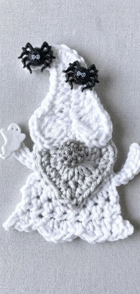 Crocheted white ghost gnome applique with a gray beard and round gray nose, two black spider buttons on the pointed cap and a small white ghost button on the left arm, photographed on plain gray fabric.