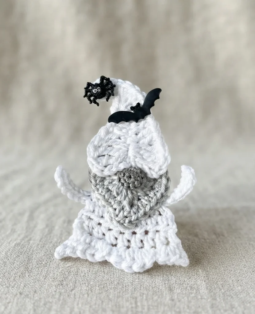 Crochet ghost gnome with a white pointed cap and two curved arms, a gray heart-shaped beard, and a white scalloped body, decorated with a small black bat and a black spider on the cap, standing on a plain beige fabric background.