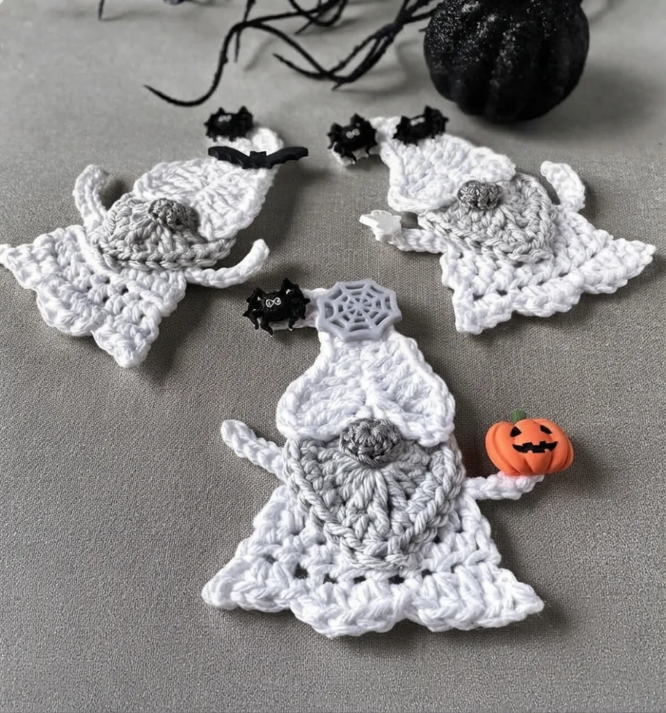 Three finished crochet ghost gnomes in white yarn with grey heart-shaped beards and pointed caps, arranged on a grey surface with a black pumpkin, black twig branches and Halloween craft buttons shaped like spiders, bats, a spider web and a